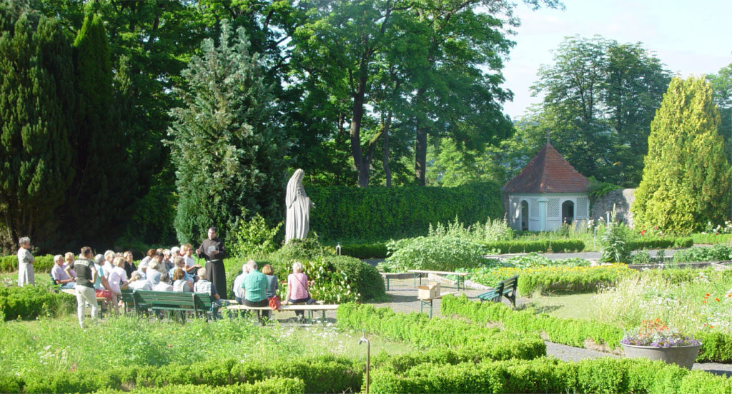 Ein Bruder des Franziskanerordens hält einen Vortrag vor einem interessierten Publikum im sommerlich grünen Garten des Klosters Frauenberg.