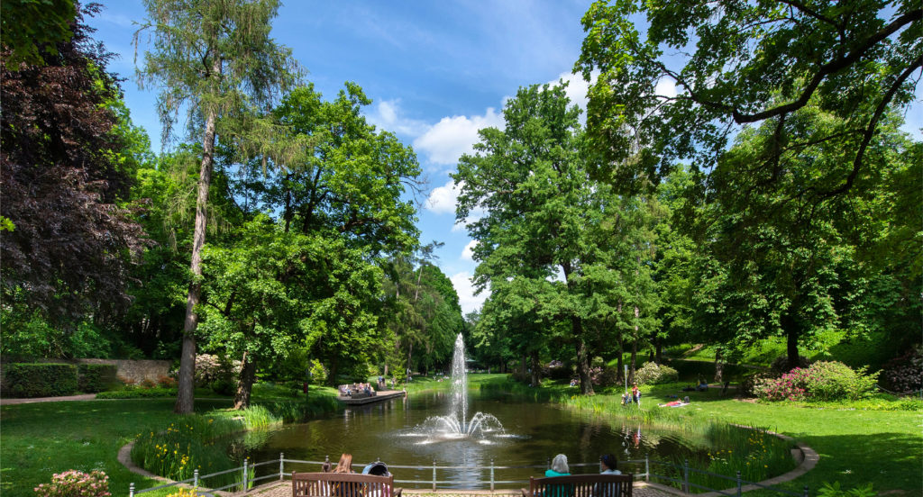 Ein Teich mit Springbrunnenanlage im landschaftlichen Teiles des Schlossgartens mit Blick auf den Holzsteg mit Sitzflächen und den Pavillon am hinteren Ende des Gartens.